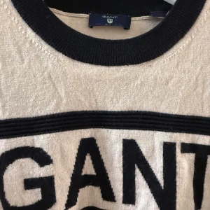 Randig beige och svart tröja från GANT - Snygg stickad tröja från GANT i beige med svarta ränder och breda muddar. Stor GANT-logga framtill, rund halsringning och långärmad modell. Perfekt för dig som gillar klassisk och sportig stil.