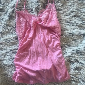 Rosa spetslinne med rosett - Supersöt rosa linne med tunna axelband och spets upptill. Linnet har en liten rosett framtill och blommiga spetsdetaljer vid bysten. Mjuk och lätt i materialet.