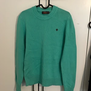 Turkos stickad tröja från Morris - Snygg turkos stickad tröja från Morris med rund halsringning och svart broderad logga på bröstet. Tröjan har ribbade muddar vid ärmslut och nederkant samt långärmad passform. Perfekt för dig som vill ha en fräsch och stilren look.