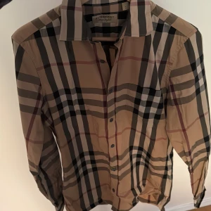 Burberry rutig skjorta i beige - Klassisk skjorta från Burberry i beige med det ikoniska rutmönstret i svart, vitt och rött. Skjortan har lång ärm, knappar framtill och en snygg krage. Perfekt för dig som gillar stilrena och tidlösa plagg med lyxig känsla.
