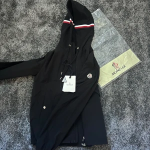 Svart vindjacka med huva från Moncler - Svart vindjacka från Moncler med huva och snörning, dekorerad med röd och vit rand på huvan. Jackan har dragkedja framtill, sidofickor med tryckknappar och Moncler-logga på bröstet. Perfekt för dig som gillar stilren och sportig look.