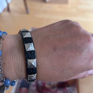 Svart läderarmband med nitar - Coolt svart armband i läder med stora fyrkantiga silvriga nitar längs hela bandet. Spännet är i silverfärg och ger en edgy vibe. Perfekt accessoar för att lyfta din outfit med lite rockig känsla.