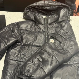 Svart Moncler pufferjacka med huva - Svart pufferjacka från Moncler med glansig finish, huva och dragkedja framtill. Jackan har två fickor med dragkedja och en Moncler-logga på ärmen. Perfekt för kalla dagar och riktigt snygg streetstil.