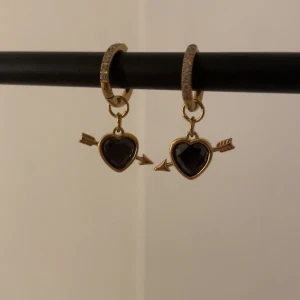 Guldörhängen med hjärtan och pil svarta - Snygga örhängen i guld med glittriga stenar på ringen och hängande svarta hjärtan som genomborras av en pil. Perfekt för dig som gillar detaljer och vill sticka ut med ett romantiskt smycke.