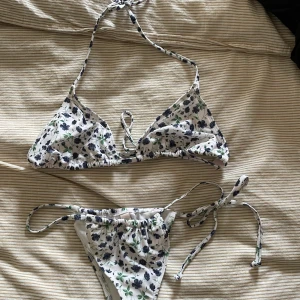 Blommig bikini från Nelly, vit/blå - Supersöt trekantsbikini från Nelly i vitt med blått och grönt blommönster. Både överdel och underdel har knytband i sidorna och runt nacken, vilket gör den enkel att justera. Perfekt för strandhäng och sommarvibbar!