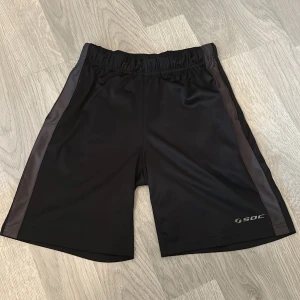 Svarta träningsshorts från SOC - Svarta träningsshorts från SOC med elastisk midja och diskreta mörkgrå sidopaneler. Tillverkade i lätt och smidig polyester som passar perfekt för sport och aktivitet. Klassisk design med logga på ena benet och bekväm passform.