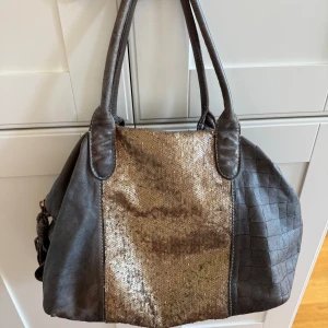 Grå handväska med paljettdetalj - Snygg brun handväska med krokopräglade sidor och breda handtag. Framsidan har en glittrig panel med guldiga paljetter som ger väskan en unik look. Tillverkad i skinn och perfekt för dig som vill sticka ut.