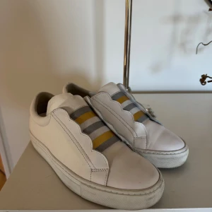 Vita sneakers med ränder - Snygga vita sneakers i skinn med elastiska band över vristen i grått och guld i storlek 38 Skorna har en vågig kant längs sidorna och en klassisk platt sula. Perfekta för dig som gillar stilrena men ändå unika detaljer. Skorna är inprincip oanvända.