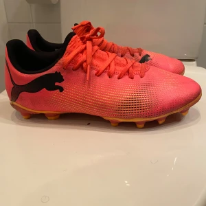 Puma fotbollsskor i neonröd och gul - Säljer ett par snygga Puma fotbollsskor i neonröd med gula detaljer och svart logga. Skorna har snörning och platt sula med dobbar för bra grepp på planen. Ovandelen är i syntetmaterial och designen är riktigt sportig och modern.