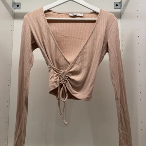 Beige ribbad omlottopp från NA-KD - Säljer en beige ribbad omlottopp från NA-KD med lång ärm och croppad passform. Toppen har djup v-ringning och snygg snörning framtill som går att justera för en cool look. Perfekt för dig som gillar trendiga detaljer och mjuka material.