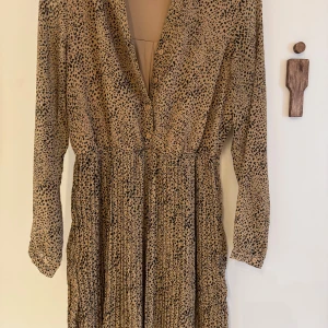 Beige långklänning med leopardmönster - Snygg beige långklänning med svart leopardmönster och v-ringning. Klänningen har långa ärmar, knappar framtill och markerad midja. 