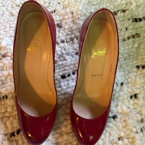 Röda klackar från Christian Louboutin - Snygga röda pumps från Christian Louboutin i lackat skinn med klassisk rund tå och hög klack. Skorna har den ikoniska röda sulan och en elegant, tidlös design som verkligen sticker ut. Perfekta för dig som vill ha ett statement-piece i garderoben.