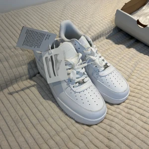 Nike Air Force 1 vita sneakers - Nike Air Force 1 sneakers i helvitt med klassisk låg siluett. Skorna har perforerad tå, snörning och rund tå. Tillverkade i skinn med vit platt sula och diskreta silverdetaljer på snörningen. Snygg och tidlös modell som passar till allt.