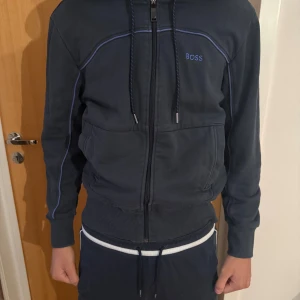 Mörkblå hoodiejacka från BOSS - Snygg mörkblå hoodiejacka från BOSS med dragkedja framtill, huva med snörning och blå kontrastsömmar. Jackan har ribbade muddar och två stora fickor framtill. Perfekt för en chill och sportig stil.