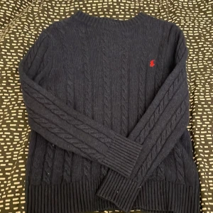 Mörkblå kabelstickad tröja Polo Ralph Lauren - Knappt använd, Stickad mörkblå tröja från Polo Ralph Lauren ser svart ut på bilden men är mörkblå med klassisk röd broderad logga på bröstet. Tröjan har rund halsringning, långa ärmar och breda ribbade muddar vid ärmslut och nederkant. Perfekt för dig som gillar stilren och tidlös design och att vara varm till vintern.