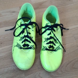 Gula adidas inomhus tränings skor med snörning - Säljer ett par riktigt coola inomhus tränings skor, från adidas i neon-gult med svarta detaljer och snörning. Skorna har platt sula och är perfekta för inomhusspel. Adidas-loggan syns tydligt baktill och på sulan. Materialet är syntet och designen är sportig och modern.