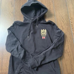 Svart hoodie från Morris  - Svart hoodie från Morris med guldfärgat emblem och brittisk flagga på bröstet. Tröjan har huva med snörning, ribbade muddar och känguruficka framtill. Perfekt för dig som gillar klassisk stil med en twist.