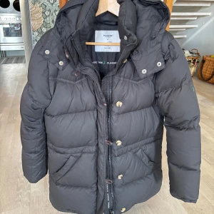 Svart dunjacka Alexandra från Boomerang  - Svart dunjacka Alexandra från Boomerang med huva och guld/bruna knappar. Jackan har dragkedja och flera fickor framtill, perfekt för kalla dagar. Stoppad med dun och har en klassisk, lite längre passform. Snygg och enkel att matcha med olika outfits. Nypris 6000kr.