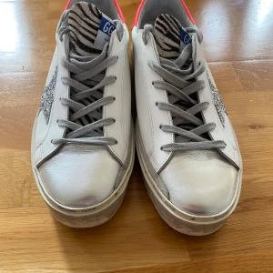 Golden Goose Hi Star sneakers med glitterstjärna - Golden Goose Hi Star sneakers i vitt skinn med grå snörning, silverfärgad tå och glittrig stjärndetalj på sidan. Hälpartiet är i stark rosa och plösen har zebramönster. Sulan är platt och har märkets logga runtom. Snygg limited edition-modell med coola detaljer. Kartong och dustbag medföljer.