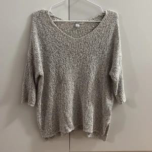 Grå stickad tröja med v-ringning - Mysig grå stickad tröja med v-ringad hals och trekvartsärmar. Tröjan har en loose passform och är perfekt för lager-på-lager under kyliga dagar. Den melerade looken ger en avslappnad och trendig vibe.