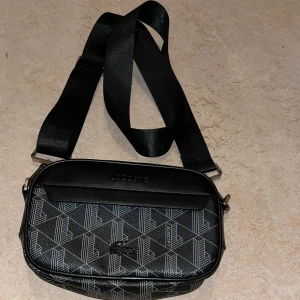 Svart axelväska från Lacoste - Snygg svart axelväska från Lacoste med geometriskt mönster och justerbar rem. Väskan har dragkedja, orange detalj på insidan och flera fack för smidig organisering. Tillverkad i syntetmaterial med stilren design och diskret Lacoste-logga framtill.