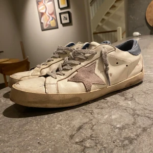 Golden goose - Säljer ett par vita sneakers från Golden Goose med grå mockadetaljer och en stor stjärna på sidan. Skorna har grå snörning, blå hälkappa och platt sula. Perfekta för dig som gillar en avslappnad och trendig stil.