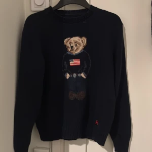 Mörkblå stickad tröja med björn från Ralph Lauren - Säljer en mörkblå stickad tröja från Ralph Lauren med ikonisk björn på bröstet. Tröjan har rund halsringning, långa ärmar och ribbade muddar. Är 180cm väger 64. Har ni övriga frågor är det bara att skriva