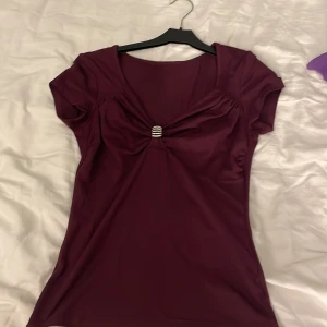 Vinröd/ lila topp med glittrig detalj - Snygg vinröd/ lila topp med korta ärmar och en glittrig silverfärgad detalj framtill som samlar tyget. Toppen har en feminin v-ringad form och är figurnära. Materialet känns mjukt och stretchigt, troligen polyester. Perfekt för att sticka ut med lite extra bling. Säljer eftersom att den inte kommer till användning. Tröjan säljs inte längre.