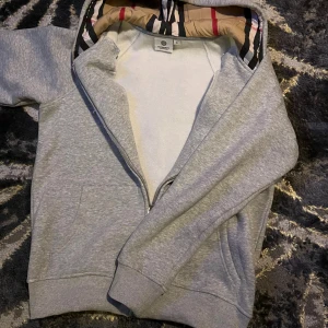 Grå Burberry hoodie med dragkedja - Säljer en grå hoodie från Burberry med klassiskt rutigt foder i huvan och broderad logga på bröstet. Tröjan har hel dragkedja, snörning i huvan och två fickor framtill. Perfekt för dig som gillar stilrena och exklusiva plagg.