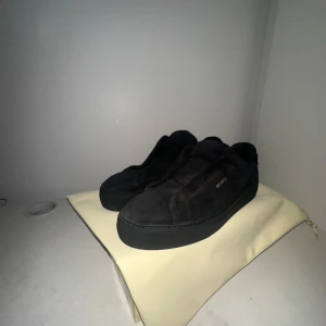 Svarta sneakers från Axel Arigato 360 lace less mocka - Snygga svarta sneakers från Axel Arigato i mjuk mocka med diskret guldfärgad logga på sidan. Skorna har en minimalistisk design med platt sula och rund tå, perfekta för en clean och stilren look. Kommer med originalpåse och låda.