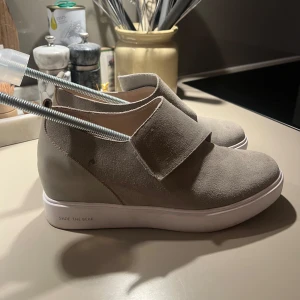 Beige sneakers från Shoe The Bear - Snygga beige sneakers från Shoe The Bear med ovandel i mocka och bred kardborreknäppning. Skorna har vit platt sula och en stilren, minimalistisk design som passar perfekt till jeans eller kjol. Bekväma och lätta att ta på tack vare kardborrebandet.