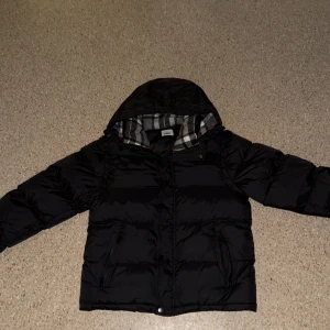 Svart pufferjacka från Burberry - Svart pufferjacka från Burberry med klassiskt rutigt foder i huvan. Jackan har dragkedja, knappar och två fickor med dragkedja framtill. Perfekt för kalla dagar med sin vadderade design och snygga detaljer.!!!Jackan är i mycket bra skick och ren. Det finns en liten reva (som syns på bilden), men den märks knappt. Därför säljer jag den till ett lågt pris. I övrigt är jackan väldigt bra, varm och bekväm.!!!!!!!!