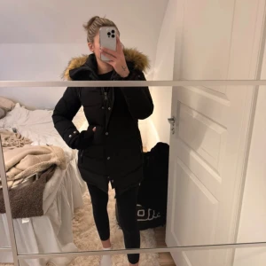 Svart lång dunjacka med fuskpäls - Svart lång dunjacka med huva och snygg fuskpäls i brunt runt kanten. Jackan har dragkedja och knappar framtill, samt fickor med dragkedja på sidorna. Jackan är från Hollies