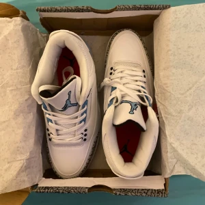 Nike Air Jordan 3 Retro Vit/Blå - Säljer ett par Nike Air Jordan 3 Retro sneakers i vitt skinn med blå detaljer och klassiskt elephant print runt tån och hälen. Skorna har snörning, rund tå och en cool blå Jumpman-logga på plösen. Perfekta för dig som vill sticka ut med en ikonisk sneaker.