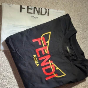 Svart Fendi t-shirt med logga - Svart t-shirt från Fendi med ikonisk Fendi Roma-logga i rött och gult framtill. T-shirten har klassisk passform, rund hals och är kortärmad. Materialet är mjuk bomull som känns skön mot huden. Perfekt för dig som vill sticka ut med en lyxig streetstyle-look.
