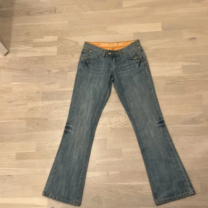 Blå bootcut jeans från cron x - Säljer ett par blå jeans från cron x med bootcut passform och klassisk femficksdesign. Jeansen har guldfärgade detaljer på bakfickorna och en snygg tvättad look. Materialet är slitstarkt denim och midjan är låg. De är köpta för 1200kr och säljer för 800kr då de är helt nya.