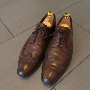 Crockett & Jones bruna brogues med snörning - Snygga bruna brogues med klassisk snörning och dekorativa perforeringar. Skorna har rund tå och platt sula, tillverkade i skinn med detaljerade mönster längs kanterna. Perfekt för dig som gillar stilrena och tidlösa skor med lite edge.