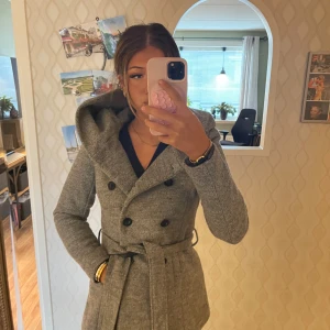 Grå kappa med huva och bälte - Säljer en snygg grå kappa med stor huva, dubbelknäppning och bälte i midjan. Jackan har långa ärmar och en klassisk, rak passform. Perfekt för dig som vill ha en stilren och mysig look under höst och vinter.