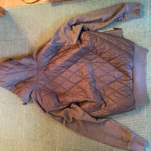 Grå cardigan Michael Kors - Snygg grå cardigan från Michael Kors med quiltad framsida och huva med snörning. Jackan har dragkedja, ribbade muddar och två fickor med knappstängning. Materialmix av polyester och stickad textil ger en modern look.