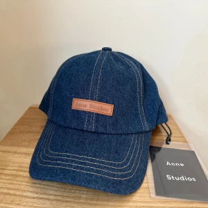 Blå denimkeps från Acne Studios - Snygg blå keps i denim från Acne Studios med kontrastsömmar och justerbar rem baktill. Framtill sitter en brun patch med Acne Studios-loggan. Perfekt accessoar för en cool och avslappnad stil. The tag was accidentally ripped off, but the kep is new. 