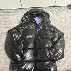 Svart pufferjacka från Polo Ralph Lauren - Snygg svart pufferjacka från Polo Ralph Lauren med glansig finish och röd broderad logga på bröstet. Jackan har huva, dragkedja framtill och flera fickor. Perfekt för kalla dagar när du vill hålla stilen.