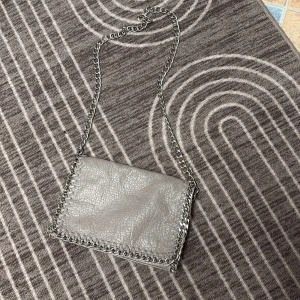 Axelväska med kedjedetaljer - Snygg grå/beige  axelväska från i skinnimitation med grov kedja som axelrem och dekorativa sömmar längs kanterna. Väskan har magnetlås, innerfack med dragkedja och ett extra fack på baksidan. Perfekt storlek för det viktigaste. Mini mini defekt sista bild. Nypris 550kr.