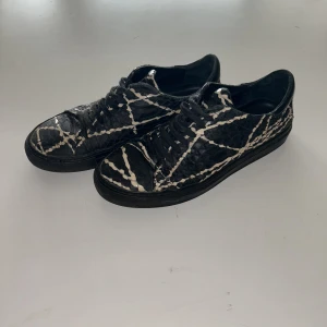 Svarta sneakers från Axel arigato - Unika svarta sneakers med vit, randig reptilmönstrad yta och snörning. Skorna har en platt sula och rund tå, tillverkade i skinn med en cool textur som sticker ut. Perfekta för dig som vill ha något annorlunda och edgy i din skogarderob.