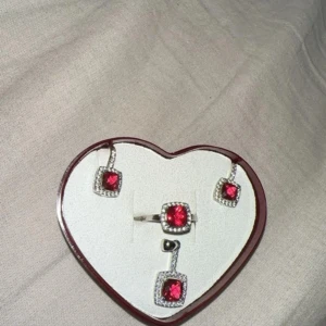 Smyckeset i silver med röda stenar - Set med ring, örhängen och hänge i äkta 925 silver. Alla delar har fyrkantiga röda stenar i mitten, omgivna av små klara stenar för extra bling. Perfekt för dig som gillar att sticka ut med färg och glittriga detaljer.           ‼️(Ring är justerbar storlek - passar alla)‼️Setet kommer i en hjärtformad ask - perfekt som present eller till dig sjalv.