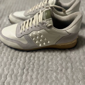 Valentino Rockrunner sneakers beige - Snygga Valentino Rockrunner sneakers i beige och grått med ikoniska nitar på sidan och hälen. Skorna har ovandel i skinn och mocka, platt svart sula och klassisk snörning. Perfekta för dig som vill ha en lyxig och trendig sneaker med unika detaljer.