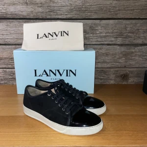 Svarta sneakers från Lanvin i mocka - Snygga svarta sneakers från Lanvin med ovandel i mocka och lackad tå. Skorna har vita platta sulor och svarta snören för en clean look. Perfekta för dig som gillar exklusiva och stilrena sneakers med lyxig känsla.