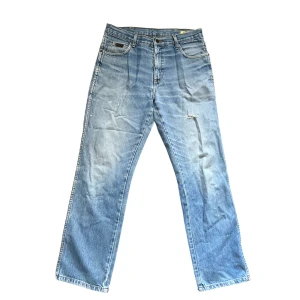 Ljusblå Wrangler jeans - Säljer ett par klassiska ljusblå jeans från Wrangler med raka ben och femficksdesign. Jeansen har snygga slitningar och ikonisk W-söm på bakfickorna samt läderpatch med logga. Perfekta för en avslappnad och tidlös stil.