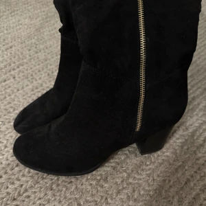 Svarta mockastövlar med klack - Snygga svarta stövlar i mocka med markerad klack och dragkedja längs sidan. Stövlarna har en klassisk rund tå och en stilren look som passar perfekt till höst- och vinteroutfits. Dragkedjan i guld ger en extra detalj till designen.