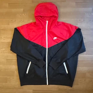 Nike röd och svart vindjacka med huva - Nike vindjacka i rött och svart med vit dragkedja och huva. ❤️🖤 Perfekt för en sportig och chill stil. Snygg färgblock-design och relaxed fit. Aldrig använd utomhus. Köptes ny för 900kr. Säljer eftersom den tyvärr inte passade i storleken