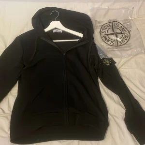 Svart hoodie från Stone Island M - Svart hoodie från Stone Island med dragkedja framtill och klassisk logga på ärmen. Helt ny utan prislapp inga defekter eller nånting. Tvättlapp, med scan så du kommer direkt till hemsidan. Hör av dig vid frågor och funderingar 